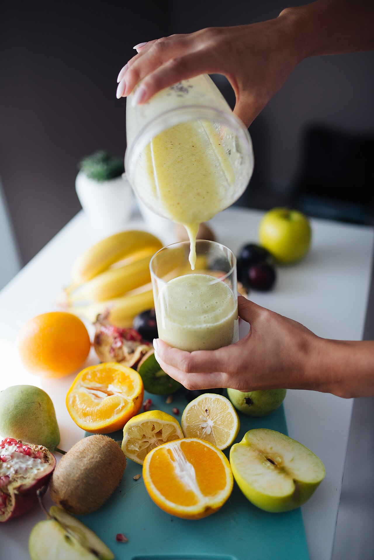 A creamy smoothie being poured from a blender into a glass surrounded by bananas, citrus, and fruit.