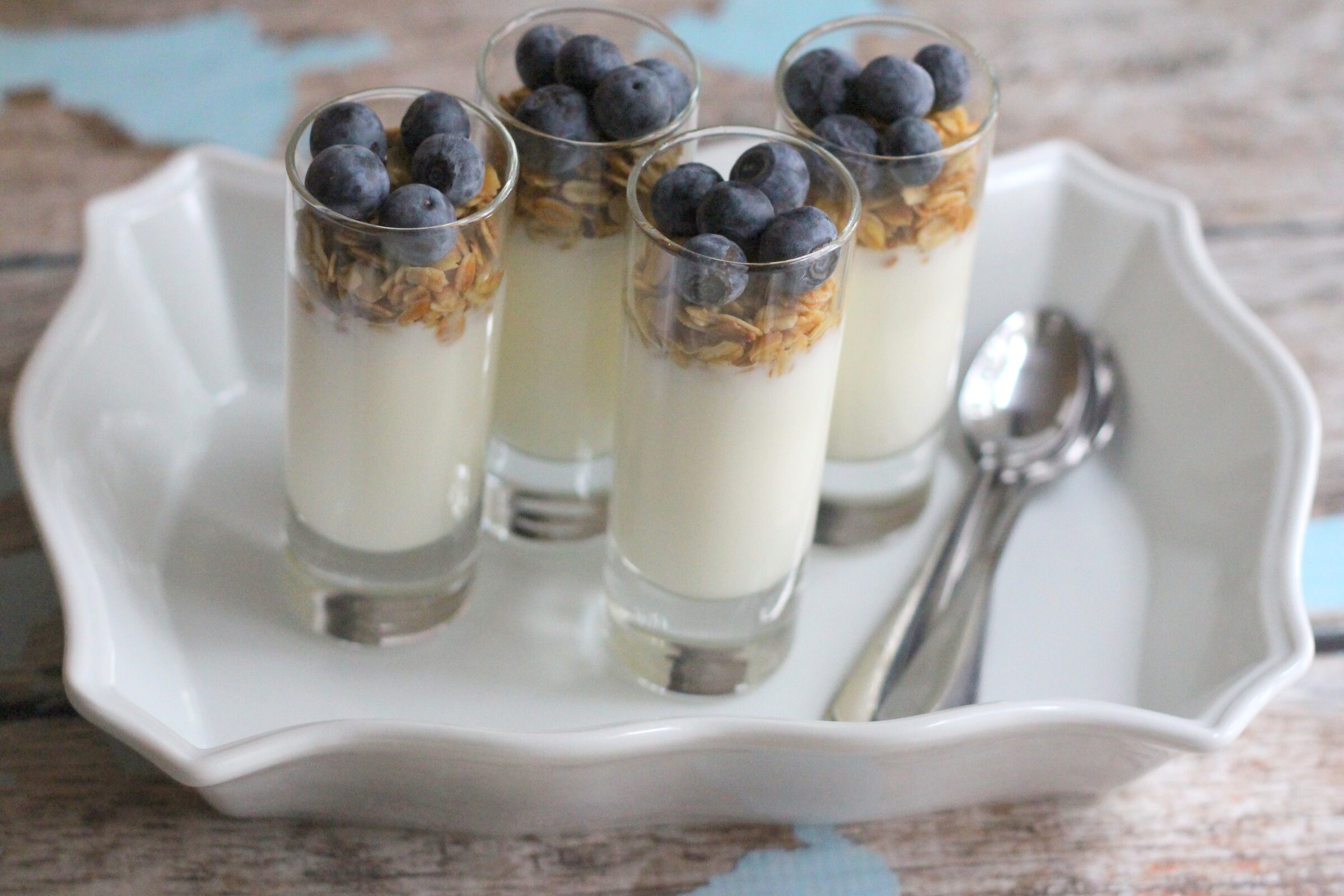 Small glasses filled with yogurt, granola, and blueberries on a white tray with silver spoons.