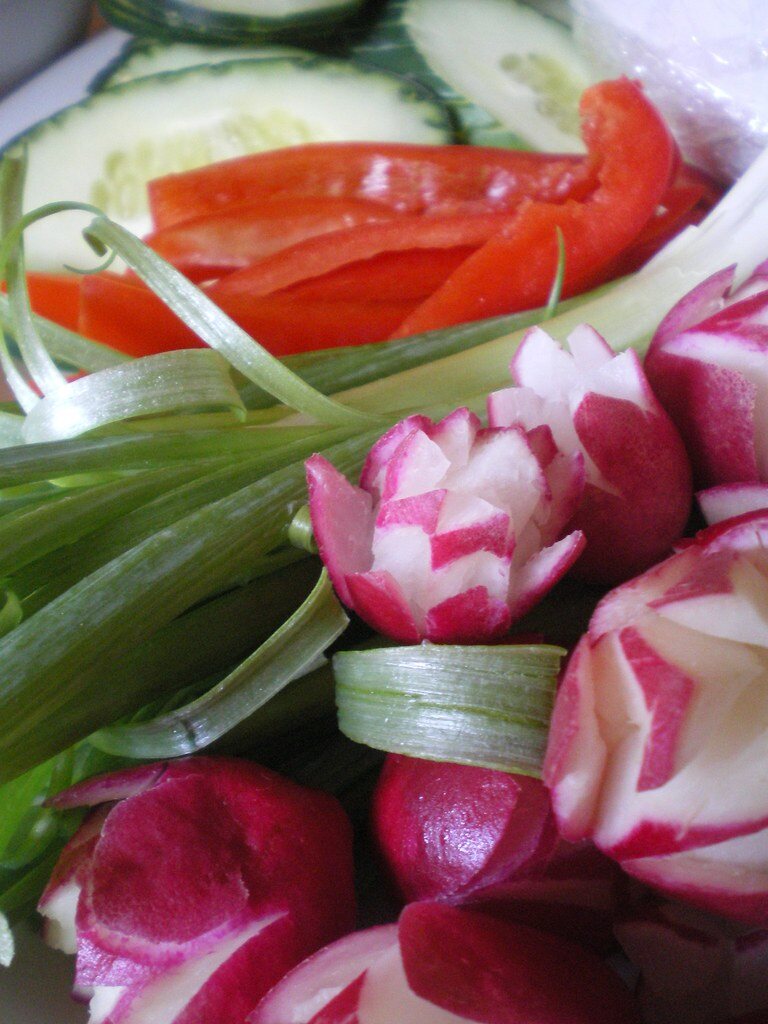 A close-up crudité platter with cucumber slices, red pepper, green onion curls, and carved radishes.
