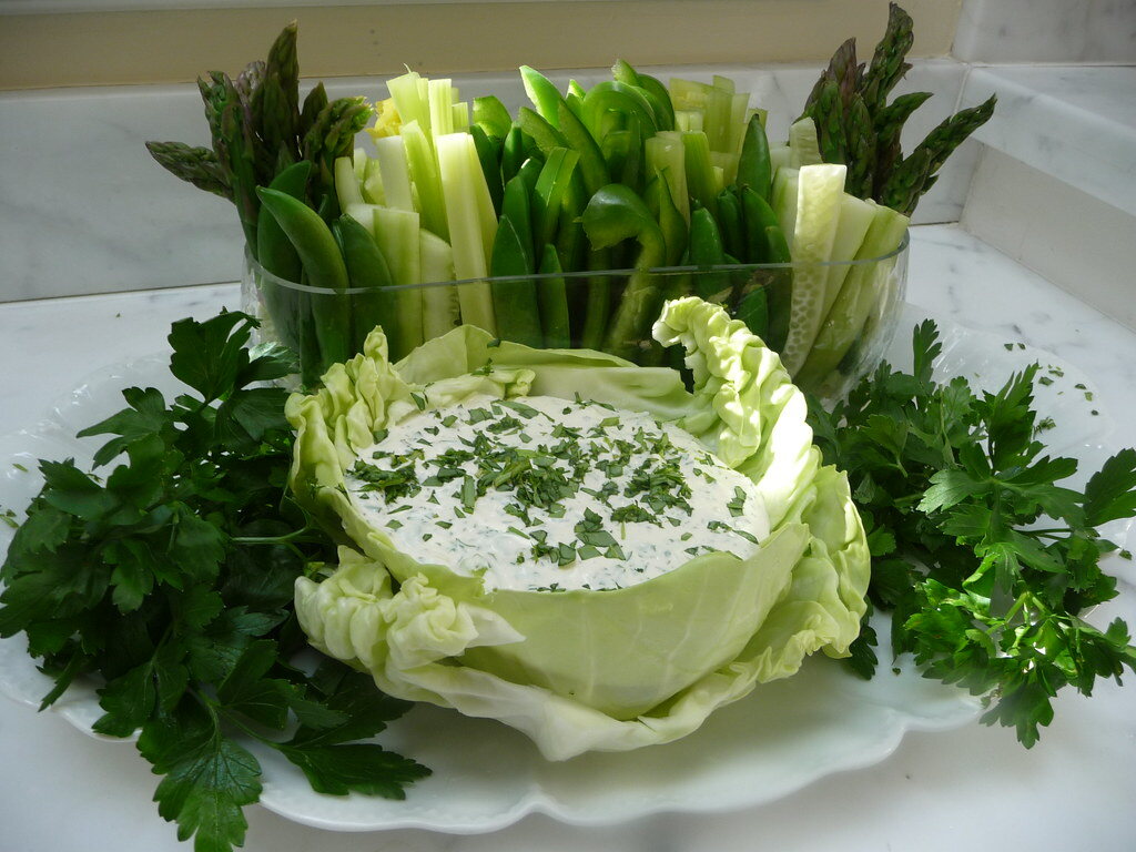 Green goddess dip served in a cabbage-leaf bowl with green vegetable sticks, asparagus, and parsley.